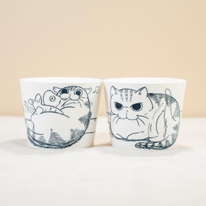 Puede incluir: Dos tazas de cerámica blanca con ilustraciones de gatos en blanco y negro. Un gato sostiene un pez y el otro gato mira al espectador.