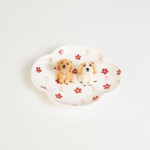 May include: A white, cloud-shaped ceramic dish with red and pink floral patterns. Two small dog figurines, one tan and one white, are placed on the dish. A decorative item for the home.