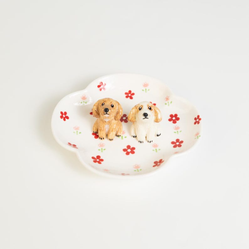 May include: A white, cloud-shaped ceramic dish with red and pink floral patterns. Two small dog figurines, one tan and one white, are placed on the dish. A decorative item for the home.