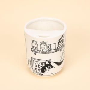 Könnte beinhalten: Ein weißer Keramik-Zahnbürstenhalter mit einer schwarz-weißen Illustration einer Katze, Regalen mit Gläsern und einem Fenster. Der Halter hat eine gewölbte Oberseite mit einem Schlitz für eine Zahnbürste.