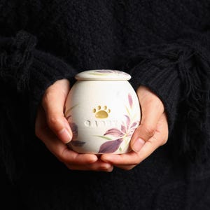 May include: White ceramic pet urn with a floral design and a paw print. The urn has the word "Gibby" inscribed on it.