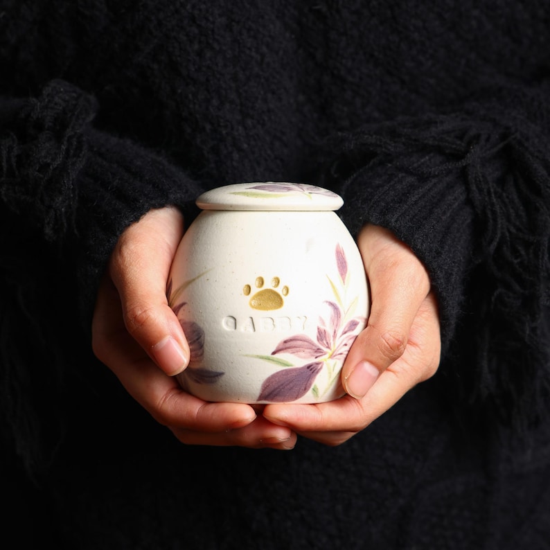 May include: White ceramic pet urn with a floral design and a paw print. The urn has the word "Gibby" inscribed on it.