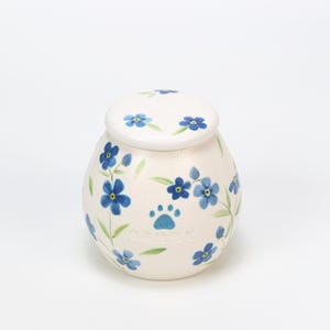 Puede incluir: Urna para mascotas de cerámica color crema con tapa, adornada con flores azules nomeolvides pintadas a mano y hojas verdes. Una huella de pata azul y el nombre "Gabby" también aparecen en la urna.