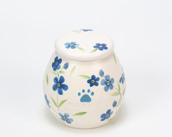 May include: A cream-colored ceramic pet urn with a lid, adorned with hand-painted blue forget-me-not flowers and green leaves. A blue paw print and the name "Gabby" are also featured on the urn.