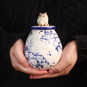 Op de afbeelding: Een witte keramische urn met blauwe bloemmotieven en de naam "LEON". Een kleine kattenfiguur staat op het blauw omrande deksel. De urn wordt met beide handen vastgehouden. Het ontwerp is traditioneel.