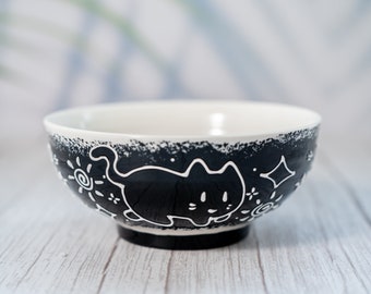 May include: A black and white ceramic bowl with a cat design. The bowl has a white interior and a black exterior with a white cat design. The cat is outlined in white and has a black interior. The bowl is sitting on a white wooden surface.