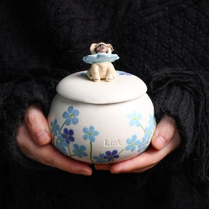 May include: A white ceramic jar with a lid, adorned with blue floral patterns and a small pug figurine on top. The jar has the word "Link" on the side. The pug wears a light blue collar.