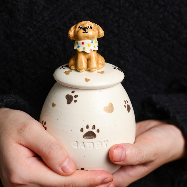 May include: A cream-coloured ceramic jar with a lid, adorned with brown paw prints and hearts. A small, brown dog figurine sits atop the lid. The jar is inscribed with the name "Gabby". The dog wears a polka-dot collar.