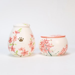 Op de afbeelding: Twee witte keramische urnen met bloemmotieven. Eén urn is eivormig met roze bloemen, groene bladeren en een pootafdruk. De andere is rond met een deksel en vergelijkbare bloemmotieven. Het woord "Alaska" is zichtbaar.