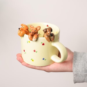 Könnte beinhalten: Eine cremefarbene Keramiktasse, verziert mit kleinen, bunten Blumenmustern. Drei braune Hundefiguren sitzen am Rand und scheinen über den Rand zu schauen. Die Tasse hat einen gebogenen Henkel und wird in einer Hand gehalten.