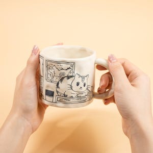 May include: A cream-colored ceramic mug featuring a hand-drawn illustration of a cat resting on a ledge. The mug has a rounded handle and a black and white drawing of a landscape scene. The cat illustration is detailed with stripes and a cute expression.