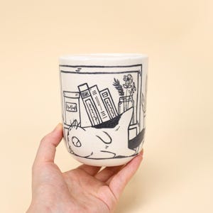 Può includere: Tazza in ceramica bianca con un'illustrazione in bianco e nero di un gatto che si rilassa davanti a una libreria. La tazza ha una forma cilindrica ed è tenuta in mano su uno sfondo color crema.