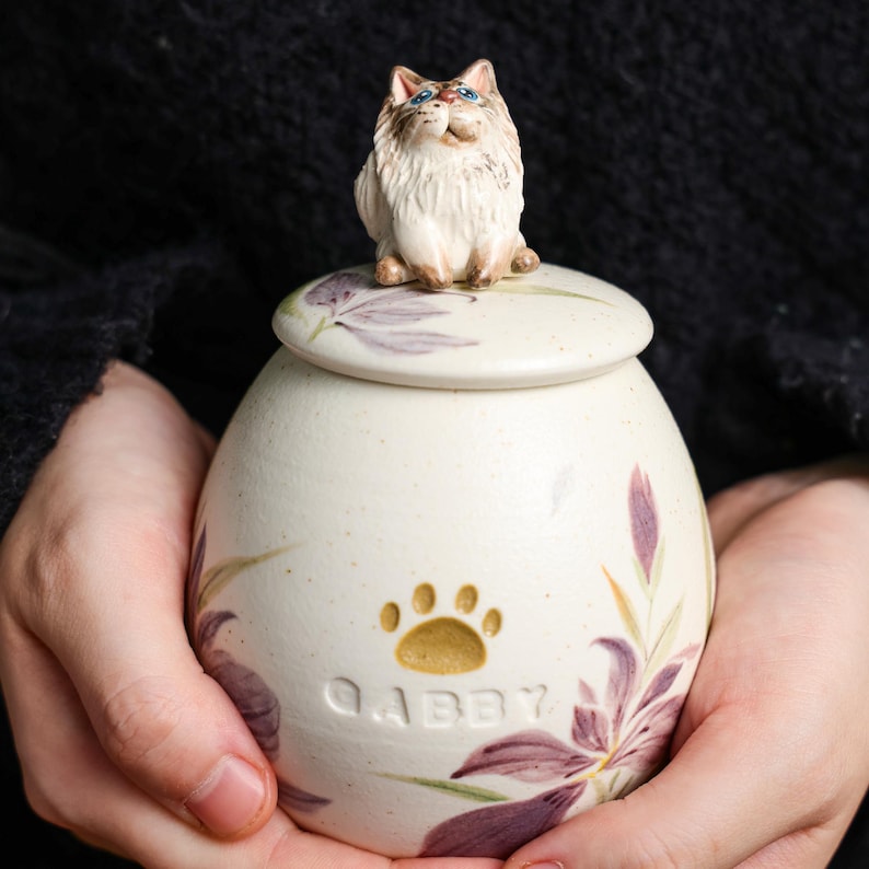May include: A cream ceramic pet urn with a lid, decorated with purple flowers and green leaves. The urn has a paw print and the name "Gabby". A small cat figurine sits on the lid. The urn is designed to hold the ashes of a pet.
