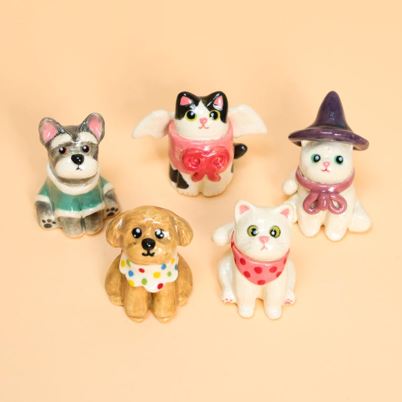 May include: Five ceramic pet figurines, including dogs and cats, are displayed. The figurines are brightly coloured and feature accessories such as scarves, hats, and wings. The background is a plain, light peach colour.