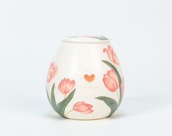 May include: A cream-colored ceramic urn with a lid, adorned with pink tulip illustrations and green leaves. A small orange heart and the word "Princess" are also featured on the urn. The urn has a speckled texture.