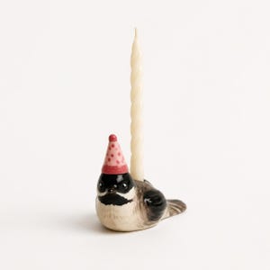 May include: A ceramic bird figurine candle holder featuring a black, white, and brown bird wearing a pink party hat with red polka dots. A tall, thin, cream-colored spiral taper candle is inserted into the top of the bird's back.