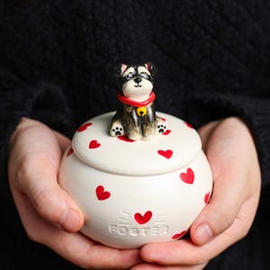 May include: A white ceramic jar with a lid, decorated with red hearts. A small dog figurine with a red collar and a yellow bell sits on top. The word "FOSTER" is printed on the jar.