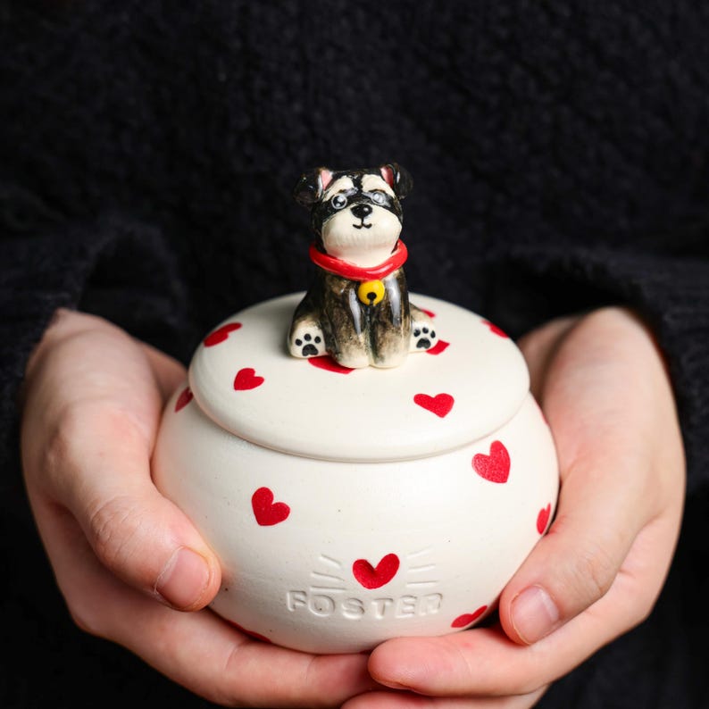 May include: A white ceramic jar with a lid, decorated with red hearts. A small dog figurine with a red collar and a yellow bell sits on top. The word "FOSTER" is printed on the jar.