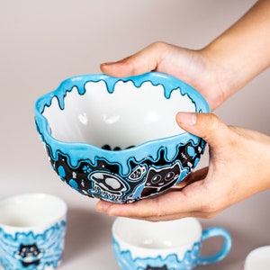 Puede incluir: Un cuenco de cerámica azul y blanco con un diseño de glaseado goteante. El cuenco presenta ilustraciones en blanco y negro de gatos, setas y otros diseños caprichosos.