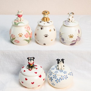 May include: A selection of ceramic pet urns, each with a unique design. The urns feature painted floral patterns, paw prints, and heart motifs. Each urn is topped with a figurine of a cat or dog. The word "Daddy" is printed on some urns.