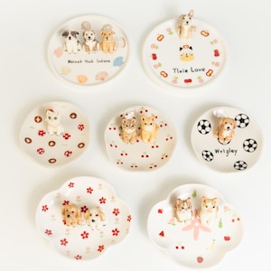 May include: A selection of decorative ceramic plates with animal figurines. The plates are white with various designs, including dogs, cats, cherries, and footballs. Some plates have text, such as "Ylvie Love" and "Wrigley."
