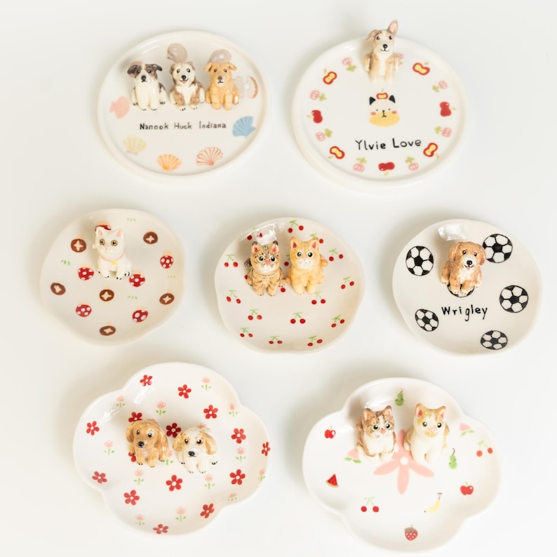 May include: A selection of decorative ceramic plates with animal figurines. The plates are white with various designs, including dogs, cats, cherries, and footballs. Some plates have text, such as "Ylvie Love" and "Wrigley."