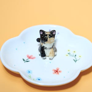 May include: A white ceramic trinket dish shaped like a four-leaf clover, decorated with small painted flowers in pink, blue, and yellow. A small, three-dimensional calico cat figurine with black, white, and tan fur sits in the center of the dish.