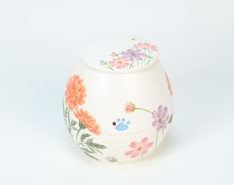 May include: Cream-colored ceramic urn with floral designs in orange, pink, and purple. The urn features a paw print and the text "Micah Anne". The lid is also decorated with flowers.