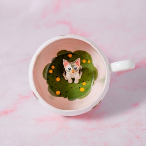 Puede incluir: Taza de cerámica blanca con un diseño caprichoso. Dentro de la taza, una pequeña y detallada figura de un gato se encuentra sobre una base verde en forma de nube con detalles amarillos. La taza tiene un asa blanca y un interior rosa.