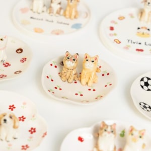 May include: A selection of small ceramic dishes, each with a unique design. The dishes feature animal figurines, including cats and dogs, and decorative patterns. The dishes are white with colourful accents and are arranged on a white surface.