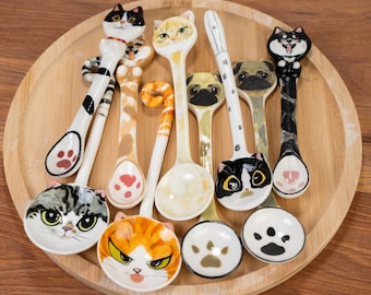 Puede incluir: Colección de cucharas de cerámica con caras de gato y pug, y huellas de patas. Las cucharas son blancas, negras, naranjas y grises, con detalles faciales. Se presentan sobre una bandeja redonda de madera.