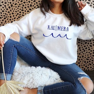 May include: White sweatshirt with blue text that reads "Kalimera" with a small fish icon and a blue wave design.