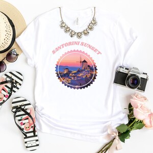 以下が含まれることがあります： 白いTシャツに、ギリシャのサントリーニ島の村の上の夕焼けを描いたピンクと白のグラフィックがプリントされています。グラフィックには風車と「Santorini Sunset」の文字が含まれています。