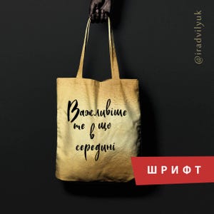 May include: A gold-coloured tote bag with two handles featuring black cursive text that reads "Важливіше те що всередині". The bag has a gradient effect, transitioning from a lighter gold tone at the top to a darker, more saturated gold at the bottom.
