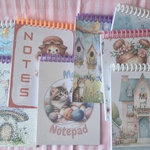 May include: A selection of spiral-bound notebooks with diverse covers. Designs feature a mushroom house, a doll, kittens with yarn, birdhouses, and a girl with flowers. The notebooks have colourful spiral bindings and text that reads "Notes", "Notepad", and "My Notebook".