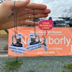 May include: Several small, white, and clear plastic air fresheners are suspended by black strings. Each air freshener features a cartoon excavator design and the text "NEIGHBORLY RENTALS (530) 362-8089". A pink sign reads "YOUR BUSINESS LOGO!"