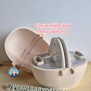 K&ouml;nnte beinhalten: Ein hellrosa Babyflaschenbeh&auml;lter mit einem umwandelbaren und abnehmbaren Deckel. Der Beh&auml;lter ist ge&ouml;ffnet und zeigt das Innere und eine graue Flasche. Der Text "Convertible and removable top" ist sichtbar.