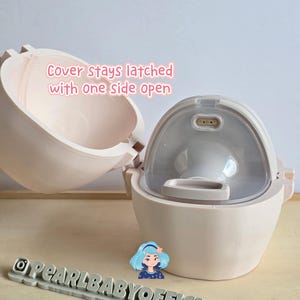 K&ouml;nnte beinhalten: Eine hellrosa Babyflasche mit einem durchsichtigen, gew&ouml;lbten Deckel. Die Flasche ist ge&ouml;ffnet und zeigt das Innere. Der Text auf dem Bild lautet "Cover stays latched with one side open." Ein graues Schild mit "@PEARLBABYOFFICIAL" befindet sich unten.
