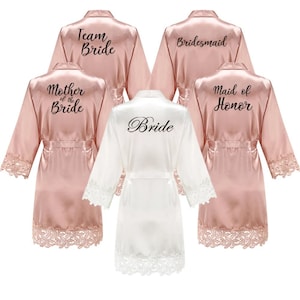 May include: A collection of satin bridal robes in shades of blush pink and white. Each robe features lace trim on the sleeves and hem. Text on the robes includes "Team Bride", "Bridesmaid", "Mother of the Bride", "Maid of Honor", and "Bride".