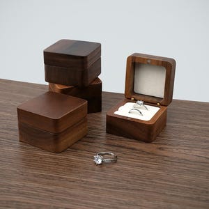 May include: Several square wooden ring boxes, one open to display two rings with clear gemstones. The boxes are a dark brown colour, and a single ring sits on the wooden surface. The boxes are made of wood.