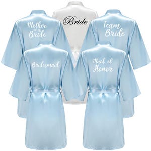 以下が含まれることがあります： ライトブルーと白のサテンブライダルパーティーローブ。「Mother of the Bride」、「Bridesmaid」、「Team Bride」、「Maid of Honor」とプリントされたライトブルーのローブと、「Bride」とプリントされた白いローブ。