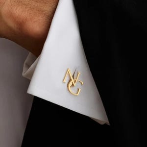 Könnte beinhalten: Nahaufnahme von goldfarbenen Manschettenknöpfen mit den Initialen "NG" auf einem weißen Hemdenärmel. Die Manschettenknöpfe sind in einer stilisierten Schriftart gehalten. Der Ärmel ist vor einem schwarzen Anzug platziert.