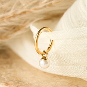 May include: A gold-colored hoop earring with a small, white pearl pendant. The earring is displayed against a backdrop of a cream-colored fabric and a textured, light-brown surface. The earring is a simple, elegant piece of jewelry.