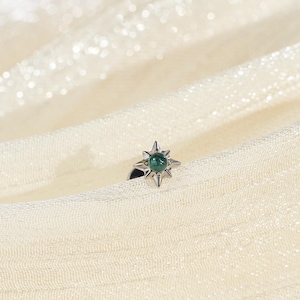 May include: A silver ring with a starburst design, featuring a central green gemstone. The ring is set against a backdrop of cream-colored, shimmering fabric, creating a soft and elegant aesthetic.