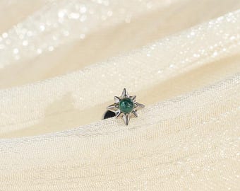 May include: A small silver-toned stud earring featuring a central round green gemstone set within a starburst-shaped metal frame with eight pointed rays extending outward from the center, creating a distinct celestial-inspired geometric design.
