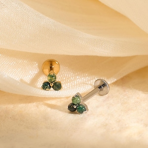 May include: Two gold-coloured stud earrings, each with a cluster of three small, round, green gemstones. One earring is upright, and the other is lying on its side. The earrings are on a cream-coloured, draped fabric background.