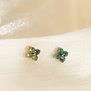 May include: A pair of small, gold-coloured flower stud earrings, each with four green gemstones. The earrings are presented on a cream-coloured, textured fabric background. The earrings have a delicate and elegant design.
