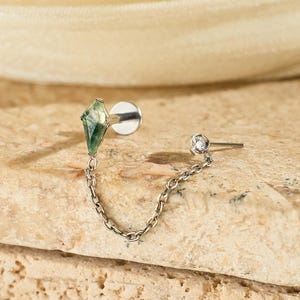 May include: A silver earring with a green, diamond-shaped stone and a small, clear gem connected by a delicate chain. The earring is set against a textured, light brown surface with a soft, off-white fabric in the background.