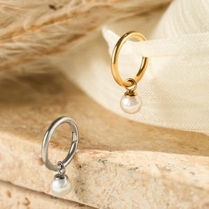 May include: Two hoop earrings, one gold-toned and one silver-toned, each with a small, dangling white pearl. The earrings are displayed on a textured, light-brown surface with a soft, white fabric in the background. A dried plant stem is also visible.