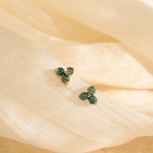 May include: Two small gold stud earrings, each featuring three round, dark green gemstones arranged in a triangular cluster. The earrings are set against a soft, cream-colored fabric background, creating a delicate and elegant aesthetic.